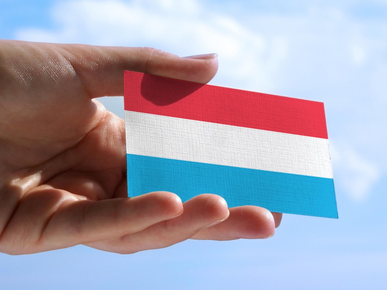 Hand mit einer Karte, auf der die luxemburgische Flagge vor einem blauen Himmel abgebildet ist.